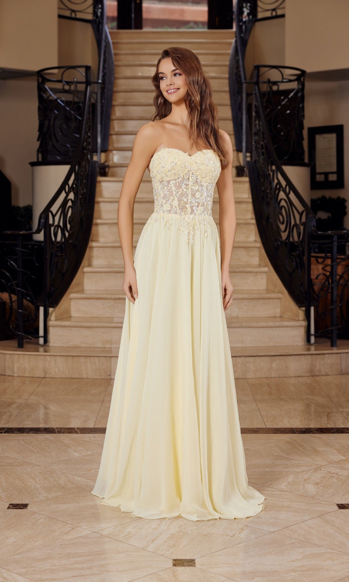 A woman stands indoors by a staircase, wearing the Nox Anabel C1617—a strapless, floor-length prom dress in light yellow with a sweetheart neckline, sheer lace bodice, and delicate floral embellishments.